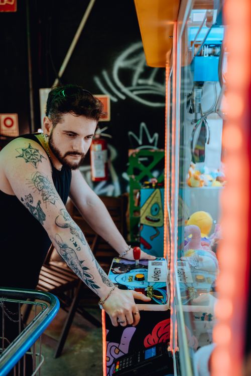 Free A tattooed young man focused on playing a claw machine in an indoor arcade with vibrant graffiti. Stock Photo