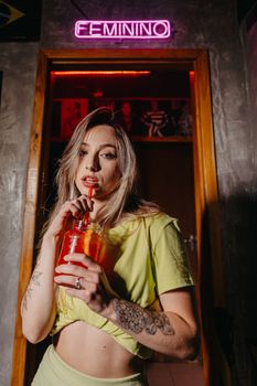 A tattooed woman in a yellow shirt sipping juice under neon light in a trendy bar.