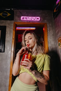 Young woman enjoying a refreshing drink with a straw, under neon lights indoors.
