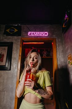 Trendy young woman sipping a cocktail under neon lights in a Brazilian nightclub.