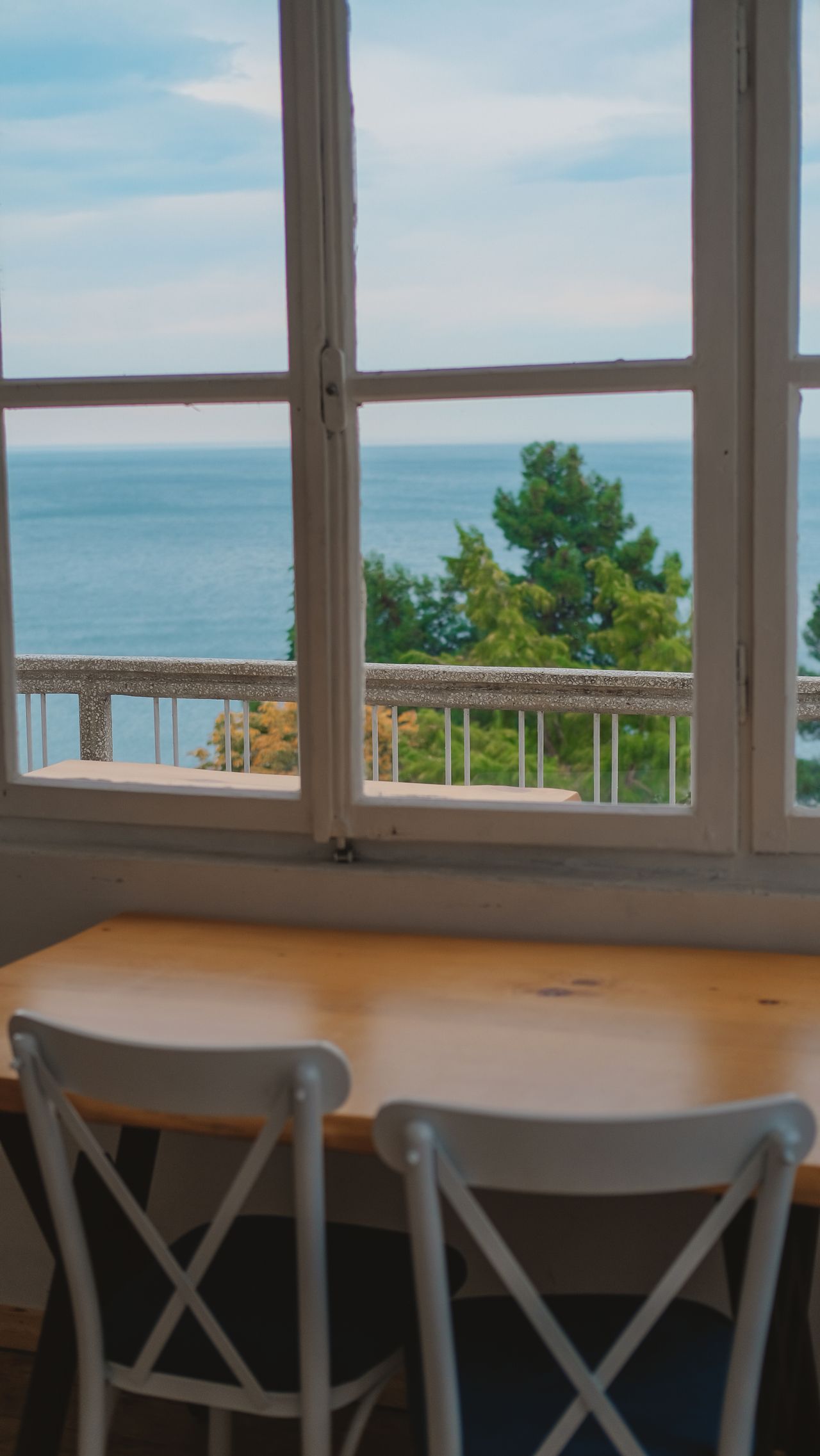 Wooden dining table with white cross-back chairs by a window overlooking water