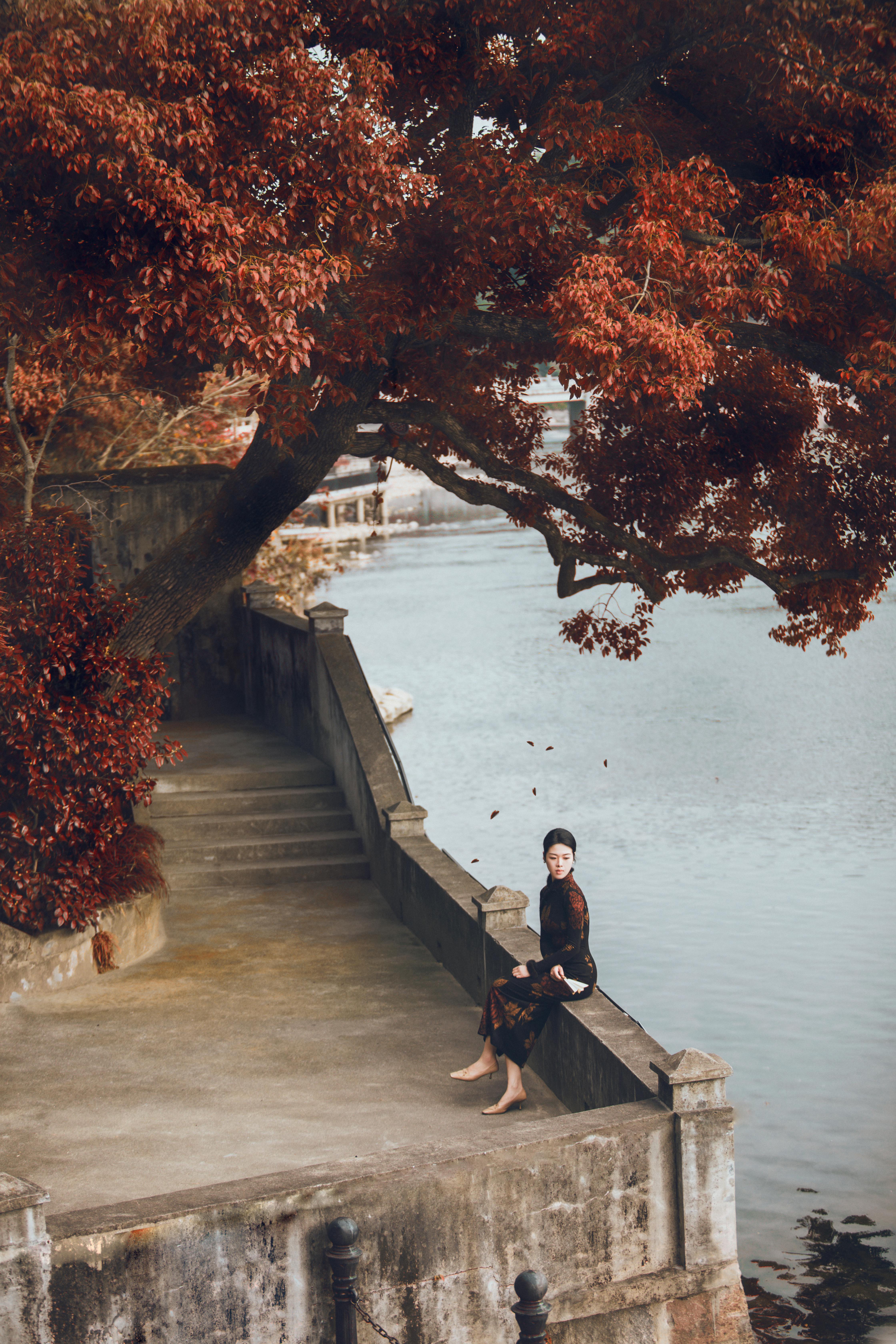 Woman in autumn setting by river, surrounded by vibrant fall foliage.
