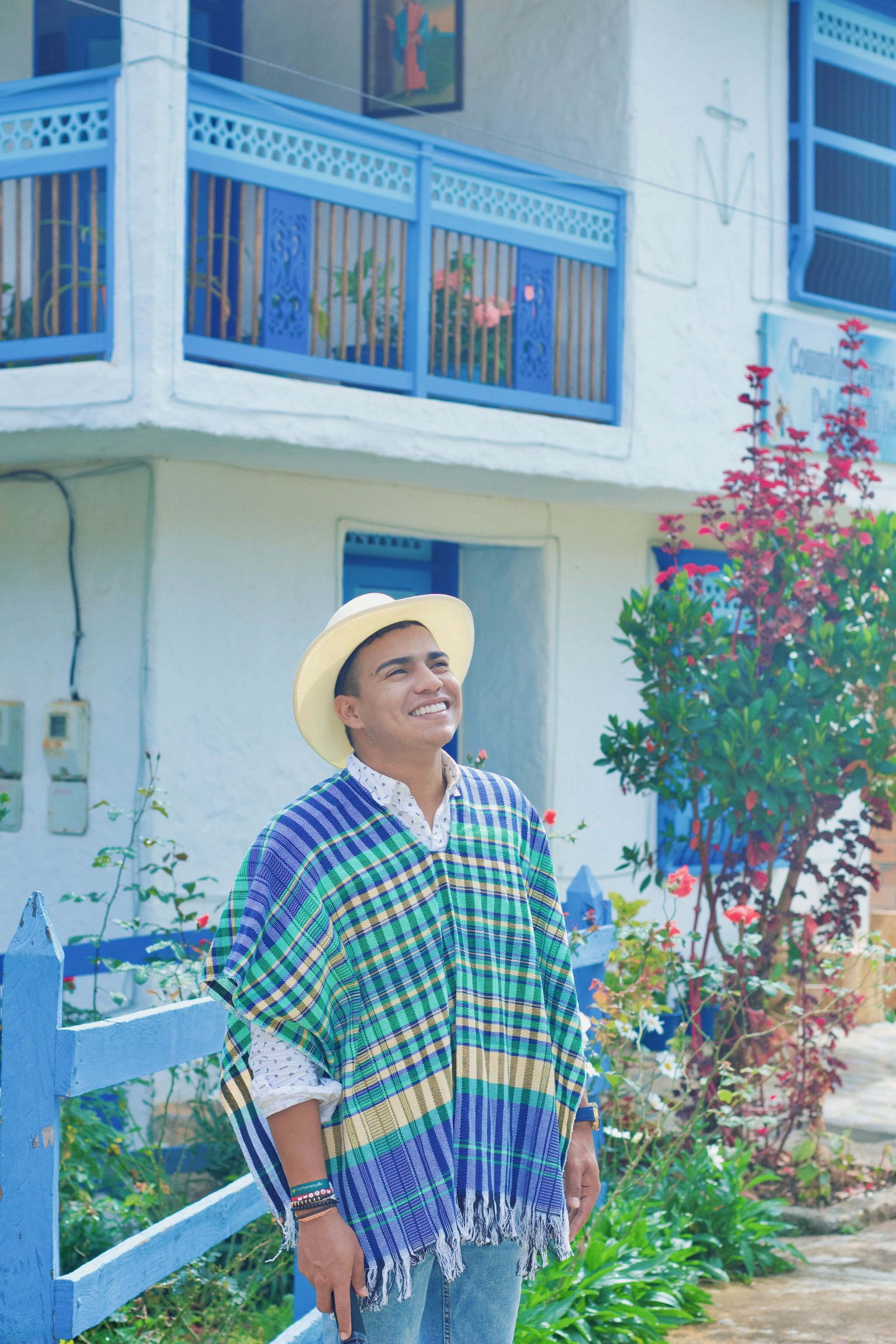 A smiling man wearing a poncho and fedora stands outdoors with vibrant surroundings, conveying joy.
