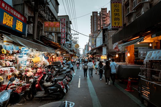 Bustling Asian city street scene with shops, pedestrians, and scooters in a lively urban shopping district.