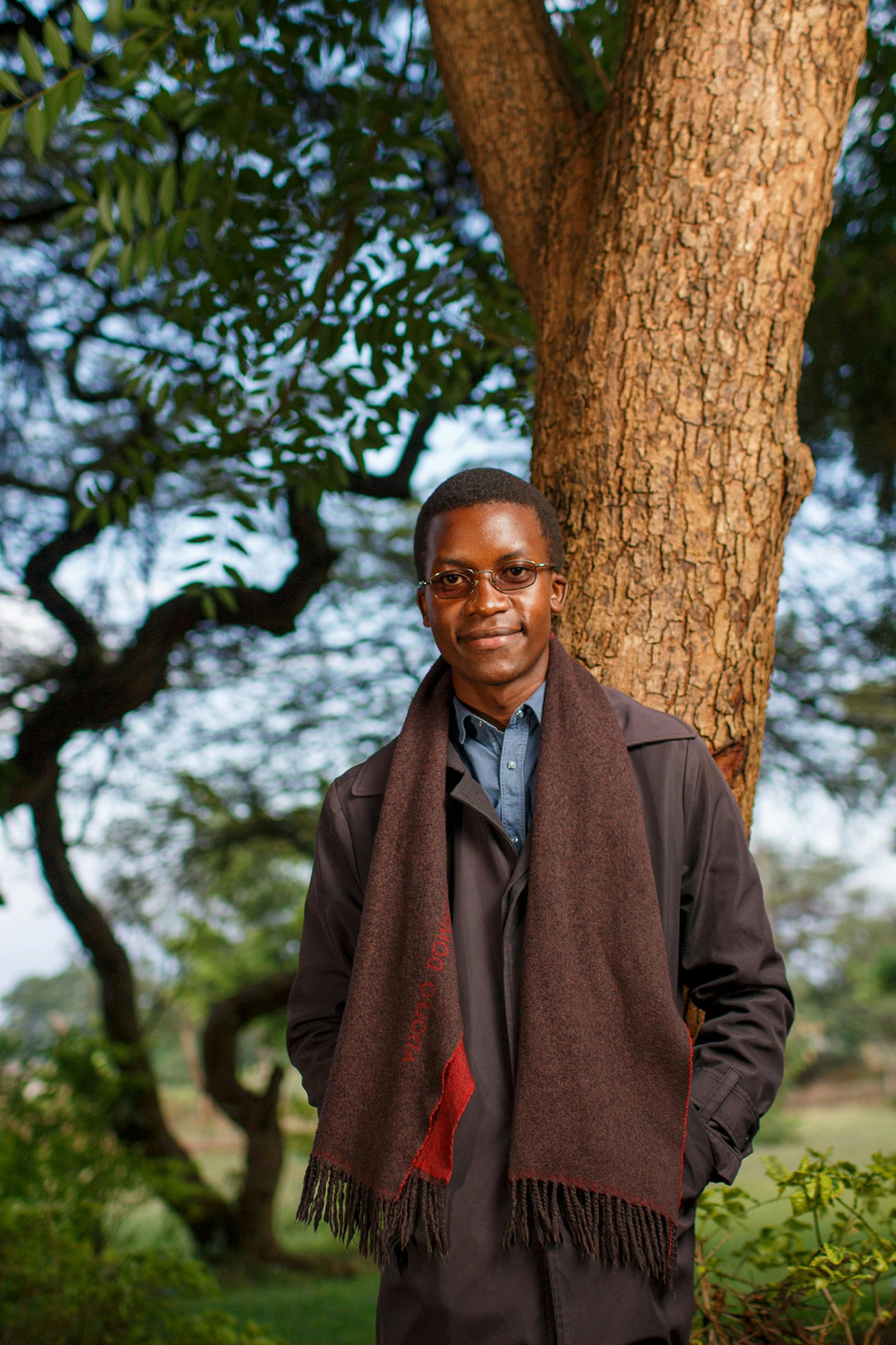 A man smiling and posing against a tree in a Lusaka park, wearing a scarf and coat.