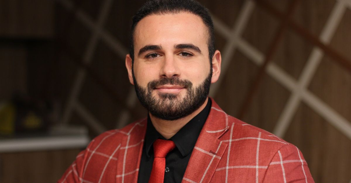 Portrait of a confident, bearded man in a red checkered suit, smiling indoors.