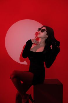 Elegant woman in a black dress and sunglasses posing against a striking red backdrop.