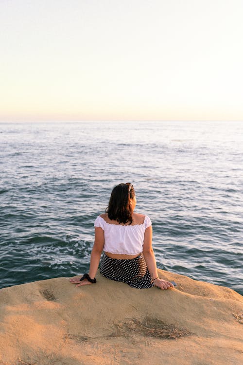 A Woman Sitting Brown Rock Formation and Looking at the Sea