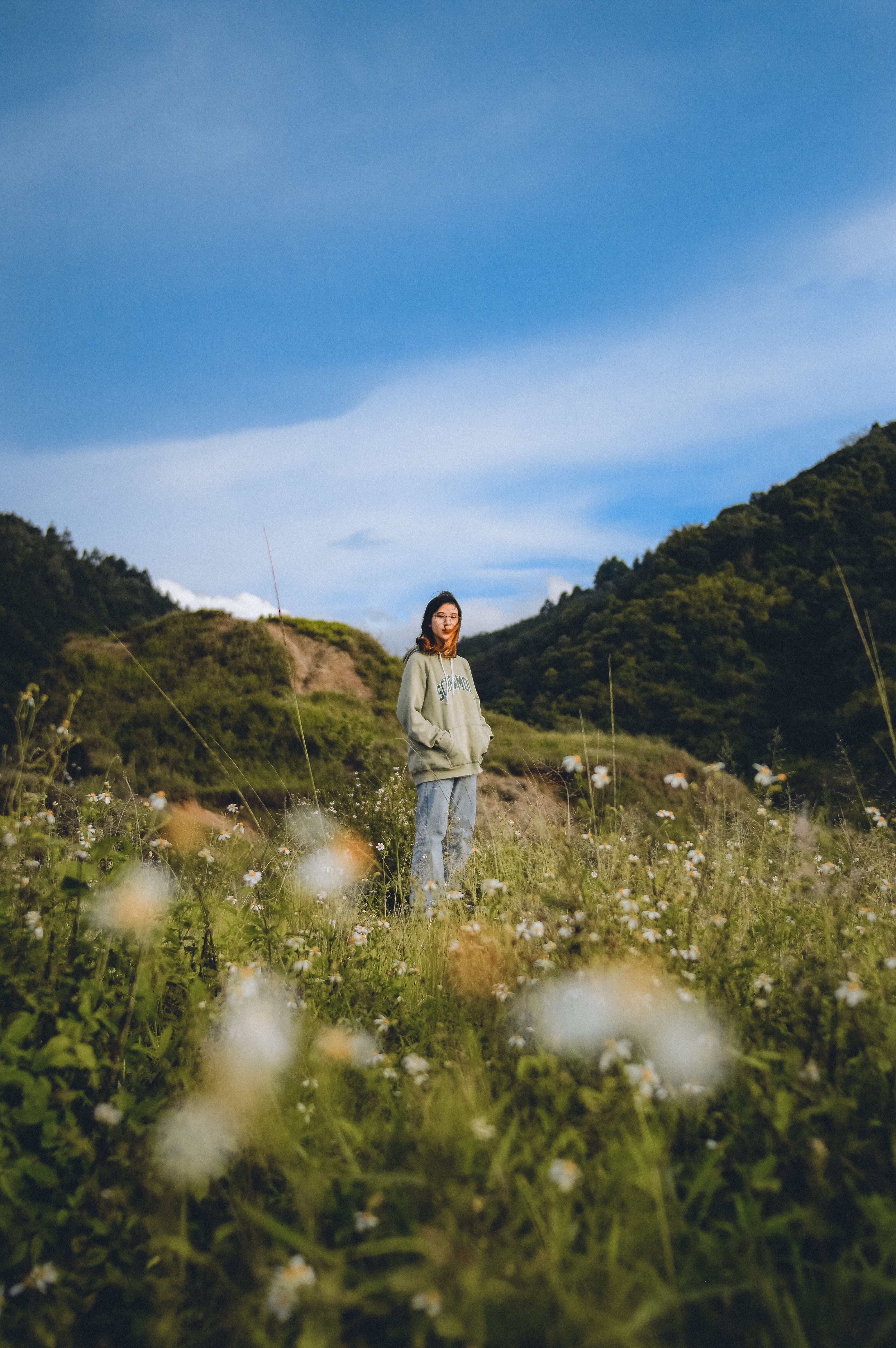 A woman stands in a blooming meadow, with scenic mountains in the background, during daylight in Indonesia.