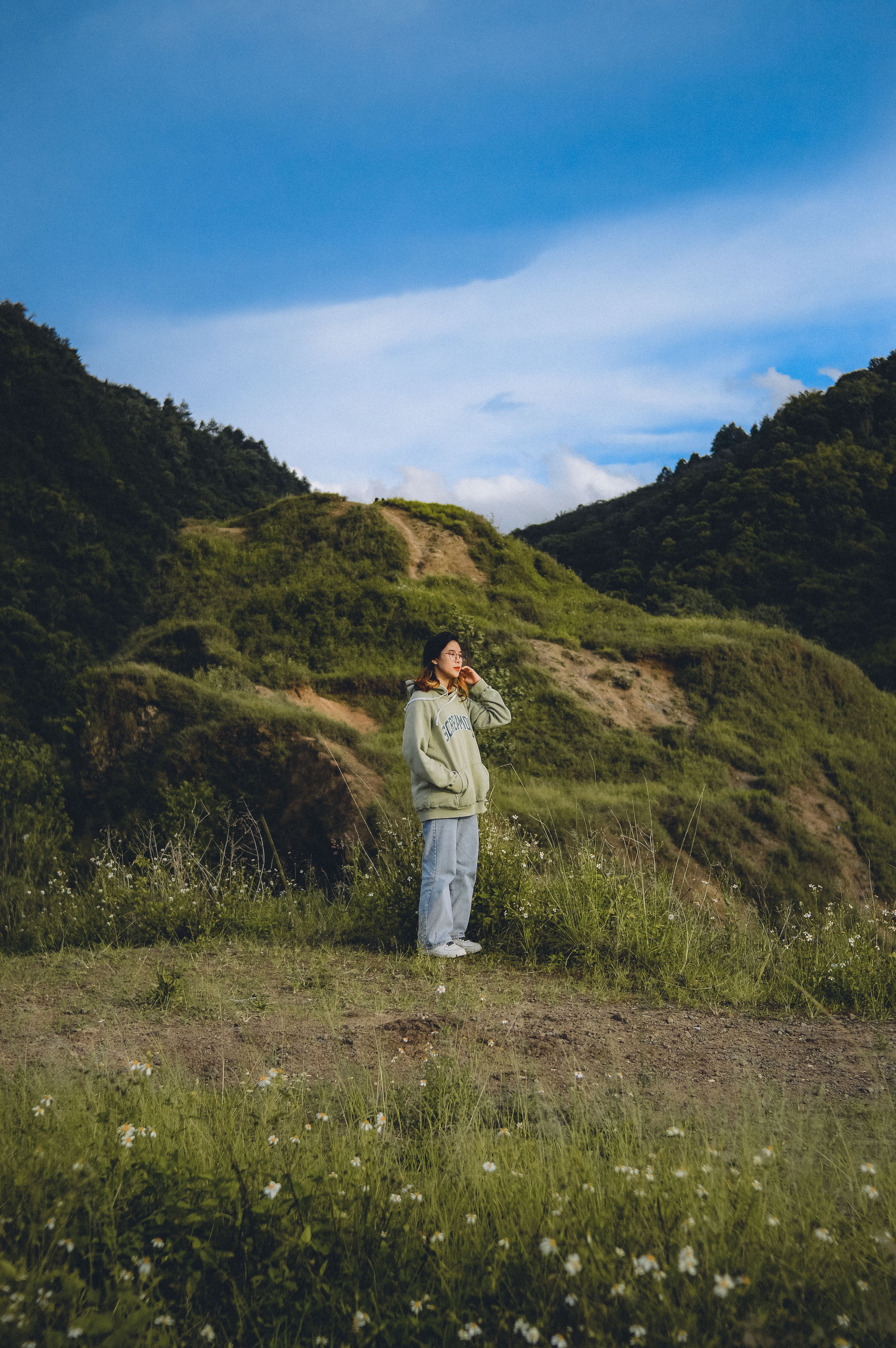 An Asian woman in a green hoodie enjoys peace in a scenic mountain landscape under a blue sky.