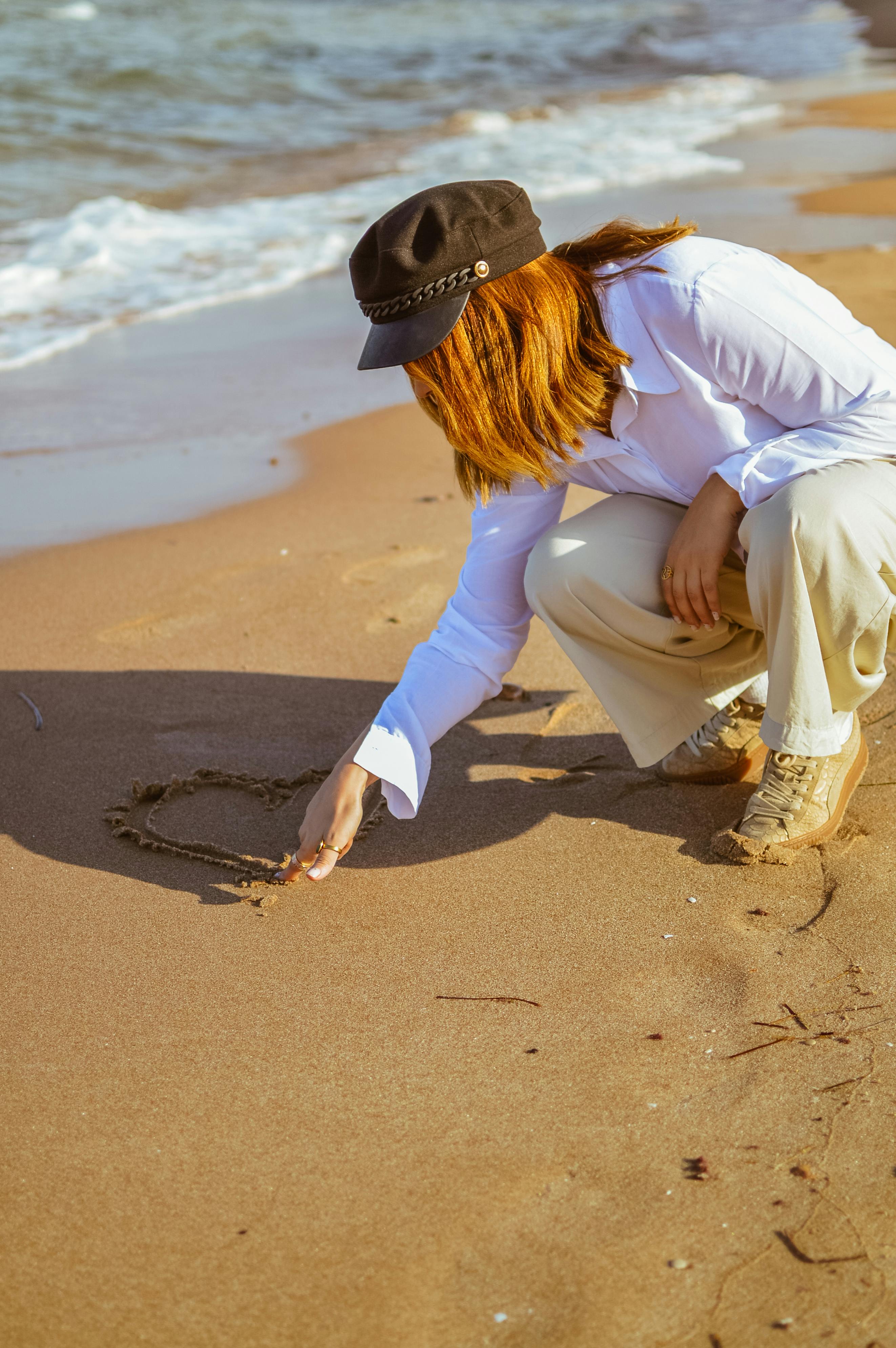 A woman crouches by the sea, drawing a heart in the sand.