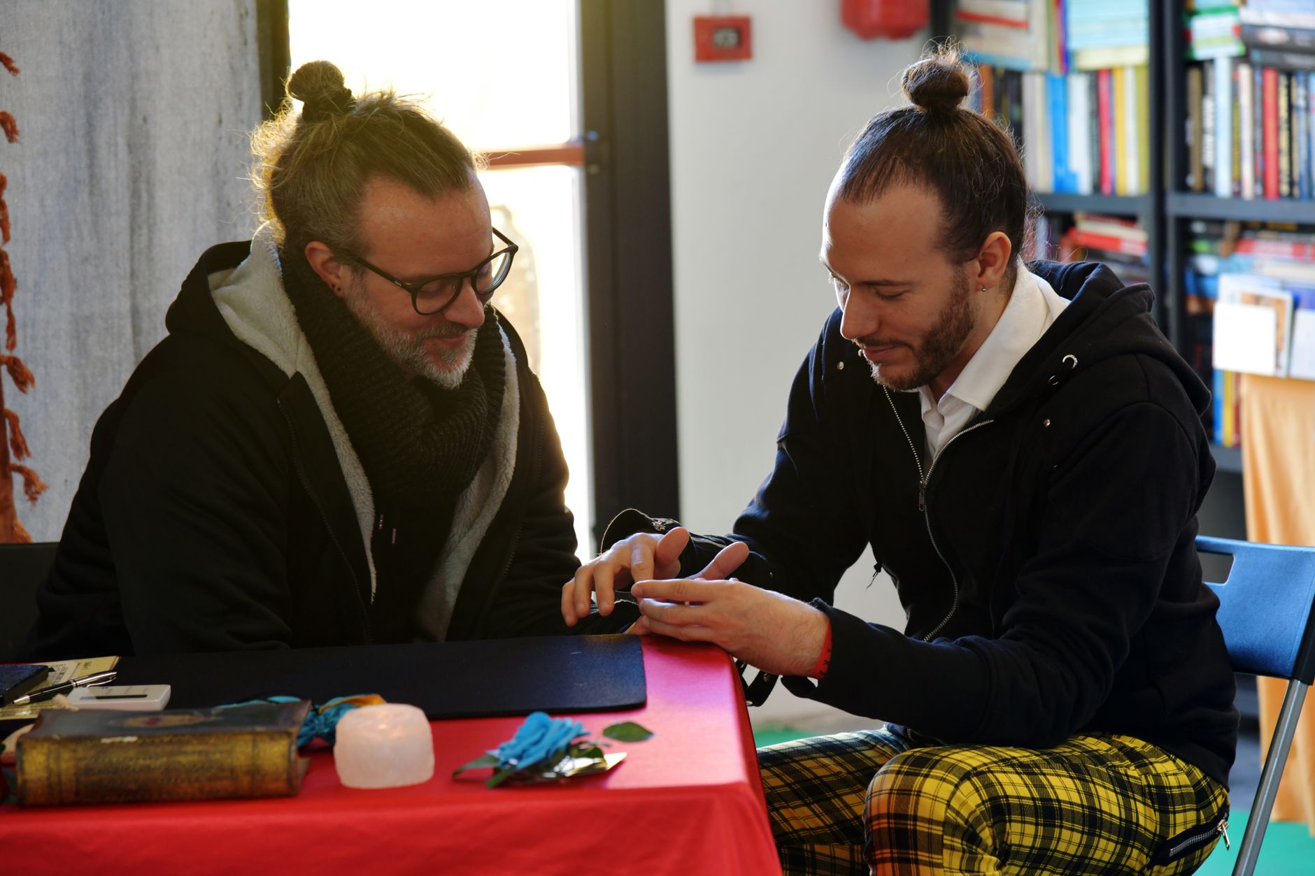 Two men engage in a casual conversation at a table, indoors, with bookshelves in the background.