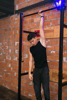 A young man in activewear exercising indoors on gym equipment in a brick-walled setting.