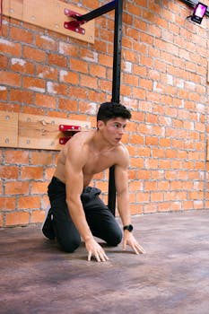 Muscular man kneeling indoors against a brick wall, focused and ready for workout.