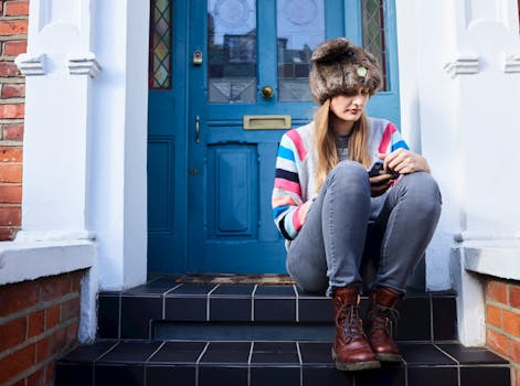 A stylish woman in a fur hat sits on a stairway, using her phone in front of a blue door.