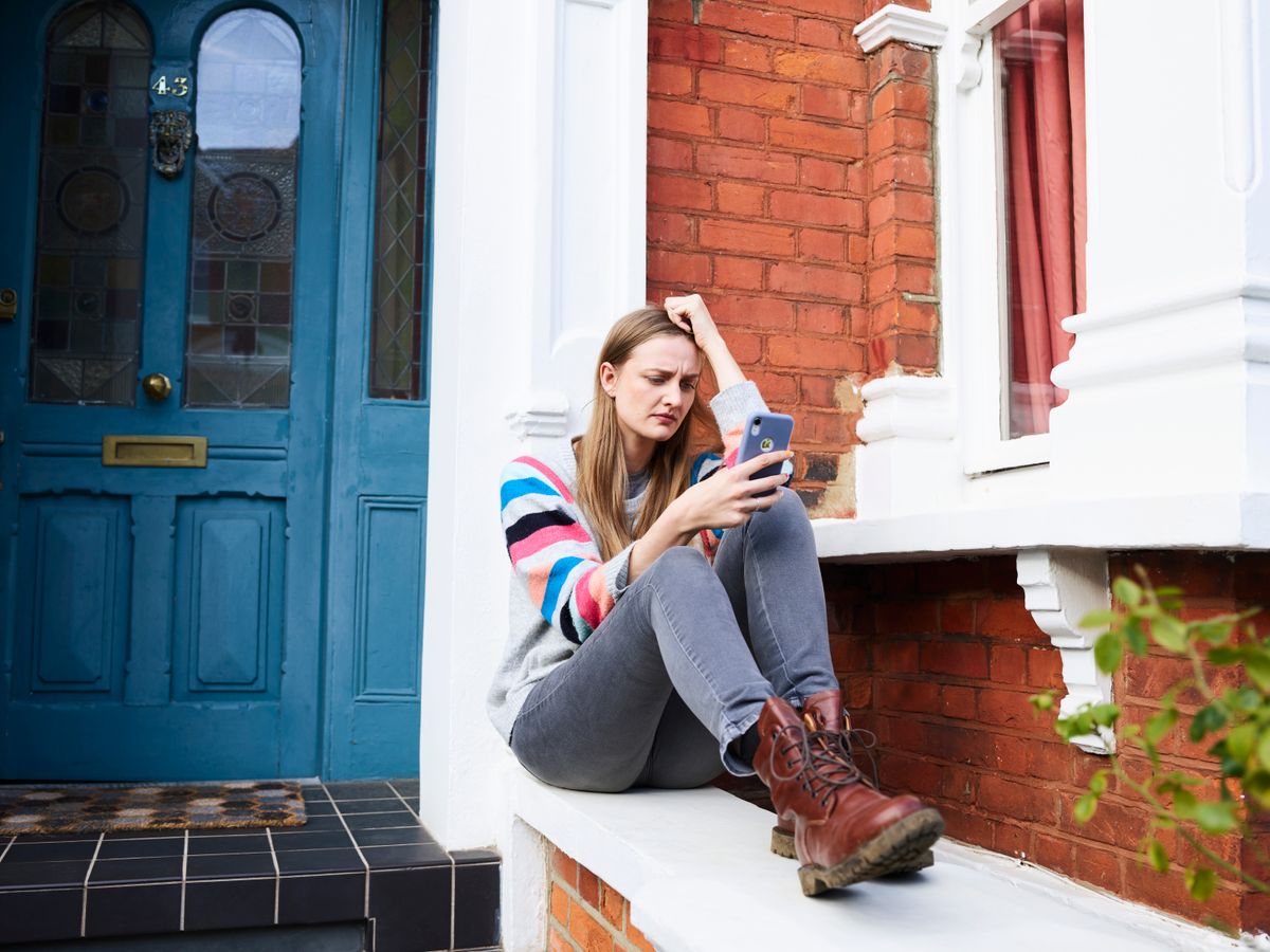 A woman sits on the white stoop of a brick rowhouse, looking at her phone after locking herself out