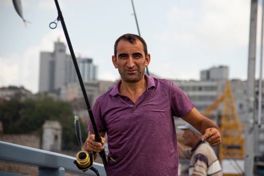A man holding a fishing rod on an urban bridge with city skyline in the background.