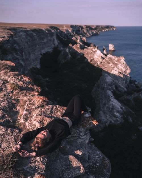 Free A woman in sportswear relaxing on a cliff overlooking a rocky coastline and sea under a clear sky. Stock Photo