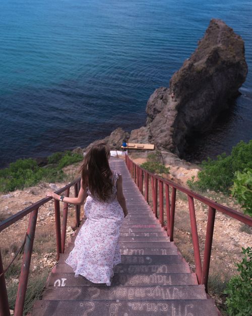 Free A woman in a white dress walks down steps towards the sea, flanked by railings and rock formations. Stock Photo
