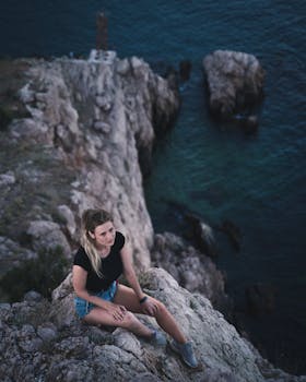 A woman sits relaxed on a cliff edge, surrounded by a scenic ocean view.
