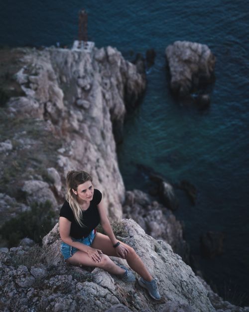 Free A woman sits relaxed on a cliff edge, surrounded by a scenic ocean view. Stock Photo