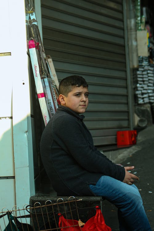 A Young Boy in Black Jacket Sitting on the Street