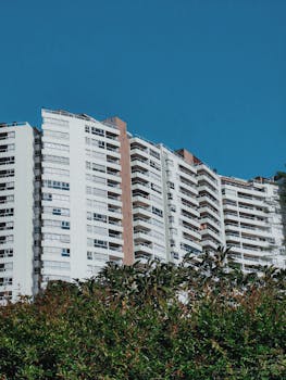 View of a modern high-rise apartment building against a clear blue sky, framed by greenery.