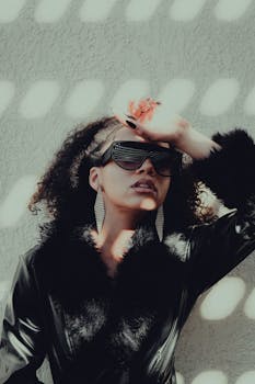 Stylish woman in sunglasses poses against a textured wall with dramatic lighting.