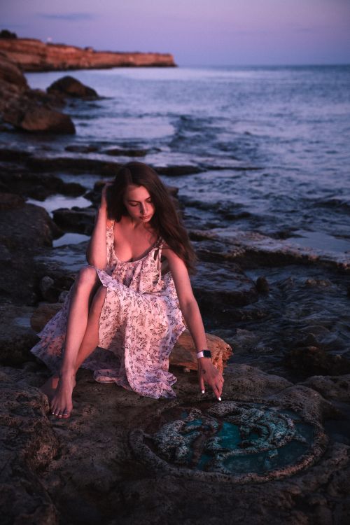 Free A serene woman in a floral dress touches rocks by the sea during twilight. Stock Photo