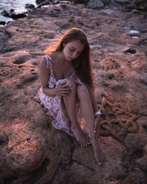 Free A young woman in a floral dress sits barefoot on intricately carved rocks by the sea. Stock Photo