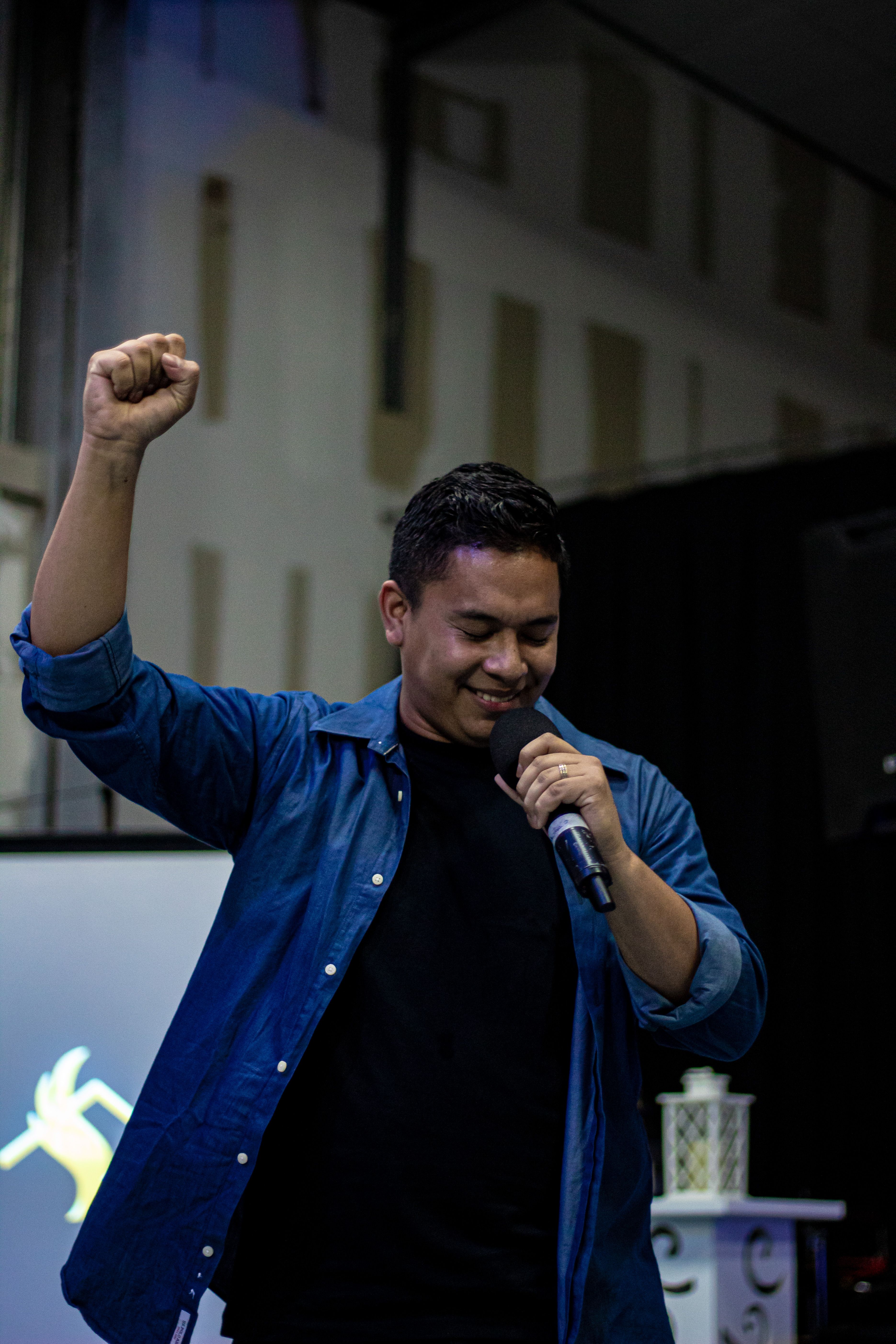 A male speaker wearing blue raises his fist in enthusiasm while speaking into a microphone indoors.