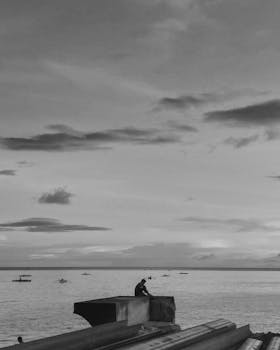 A person sits alone on a sea wall overlooking a calm ocean under a cloudy sky.