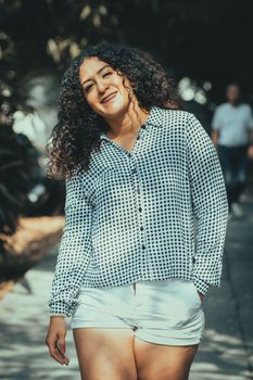 Portrait of a smiling woman with curly hair in sunlight, standing outdoors in casual attire.