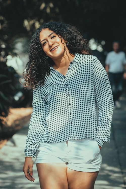 Free Portrait of a smiling woman with curly hair in sunlight, standing outdoors in casual attire. Stock Photo