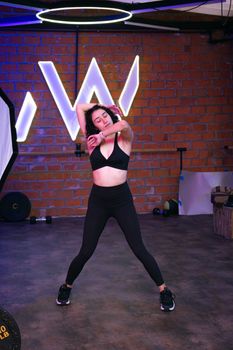 A woman in activewear stretches in a modern gym with neon lights and brick wall.