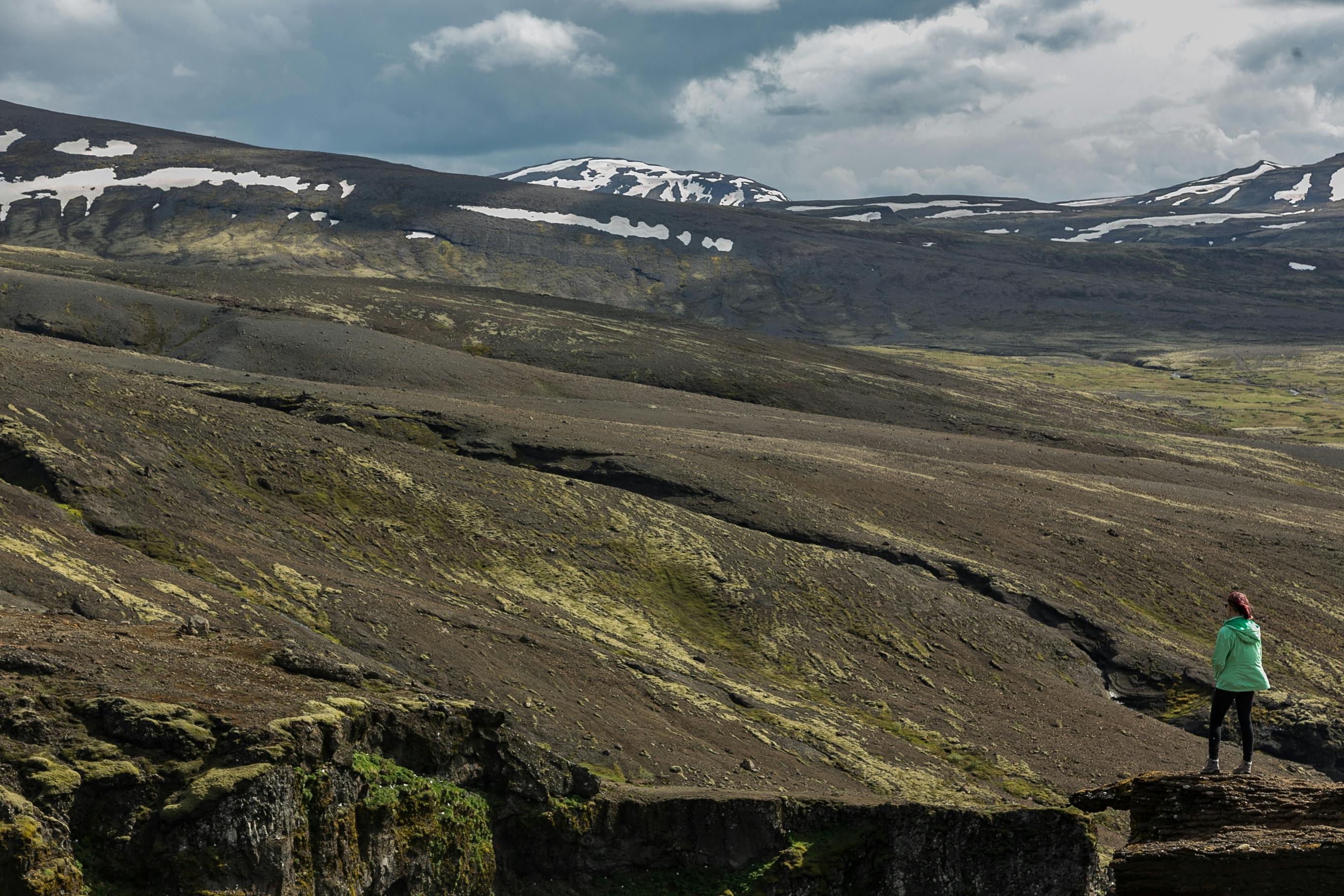 一名身穿绿色夹克的妇女站在悬崖上，俯瞰着冰岛白雪皑皑的山峰