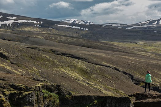 一名身穿绿色夹克的妇女站在悬崖上，俯瞰着冰岛白雪皑皑的山峰