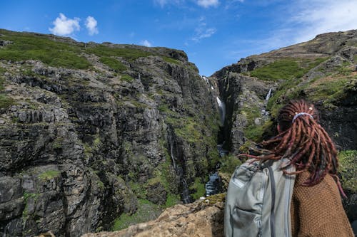 Person Wearing Brown Sweater and Grey Backpack With Brown Hair Dreadlocks on Top of Mountain Overlooking Waterfalls