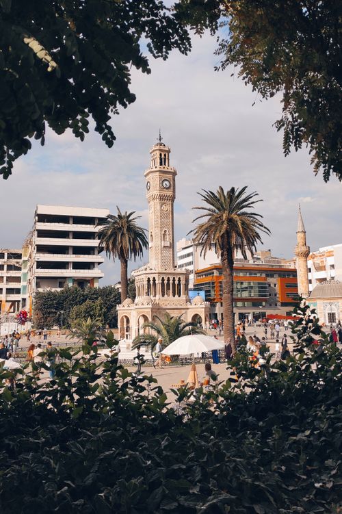 Free View of a bustling urban square with a historic clock tower surrounded by palm trees and people. Stock Photo