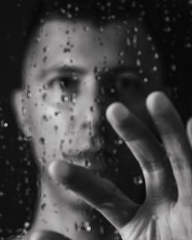 A dramatic black and white portrait of a man reaching out to a wet glass surface.