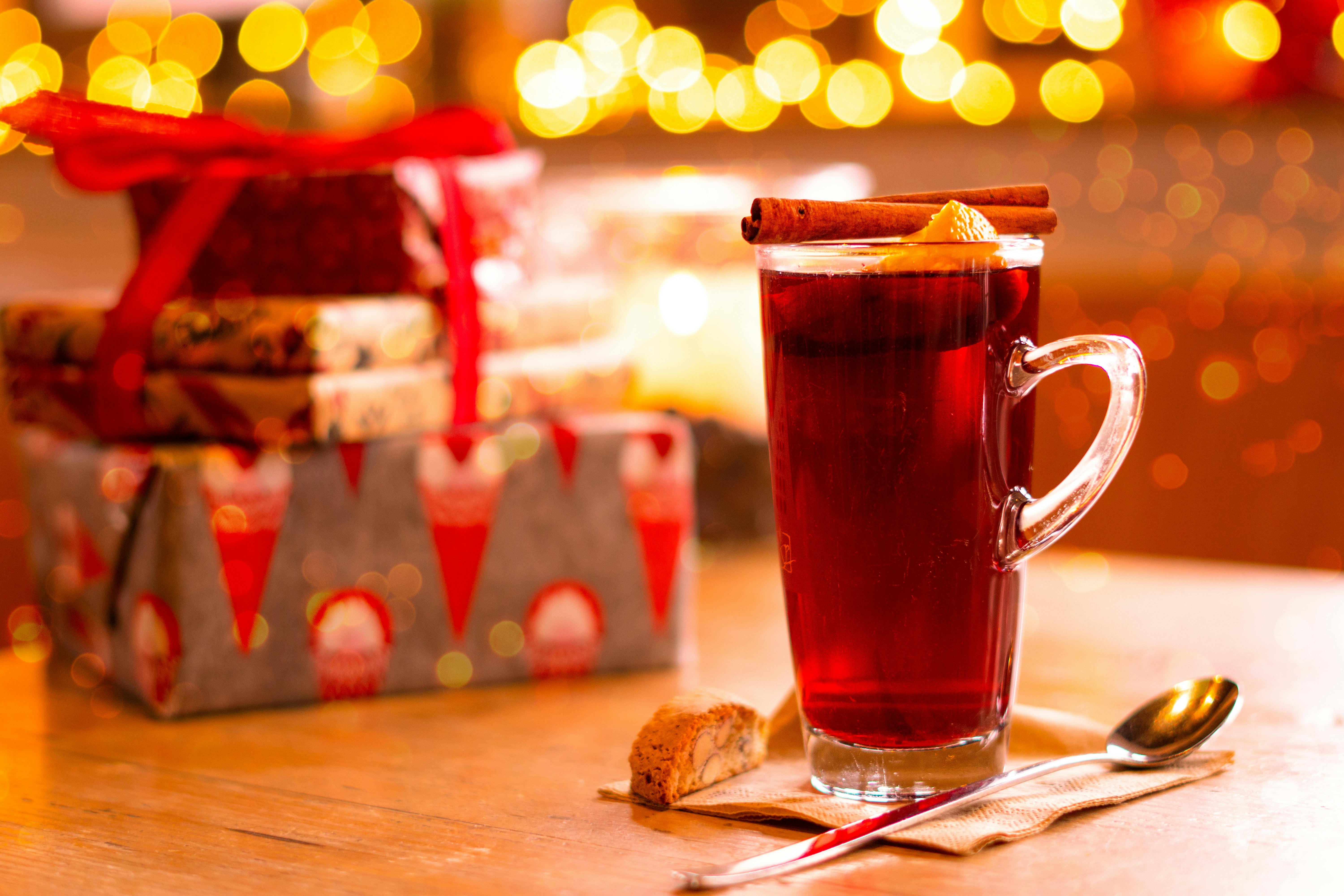 A hot glass of winter tea with cinnamon sticks, surrounded by Christmas gifts and festive lights.
