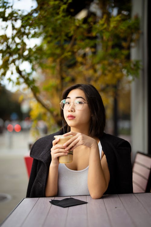 Young Woman Sitting at a Table and Holding a Cup of Iced Coffee 