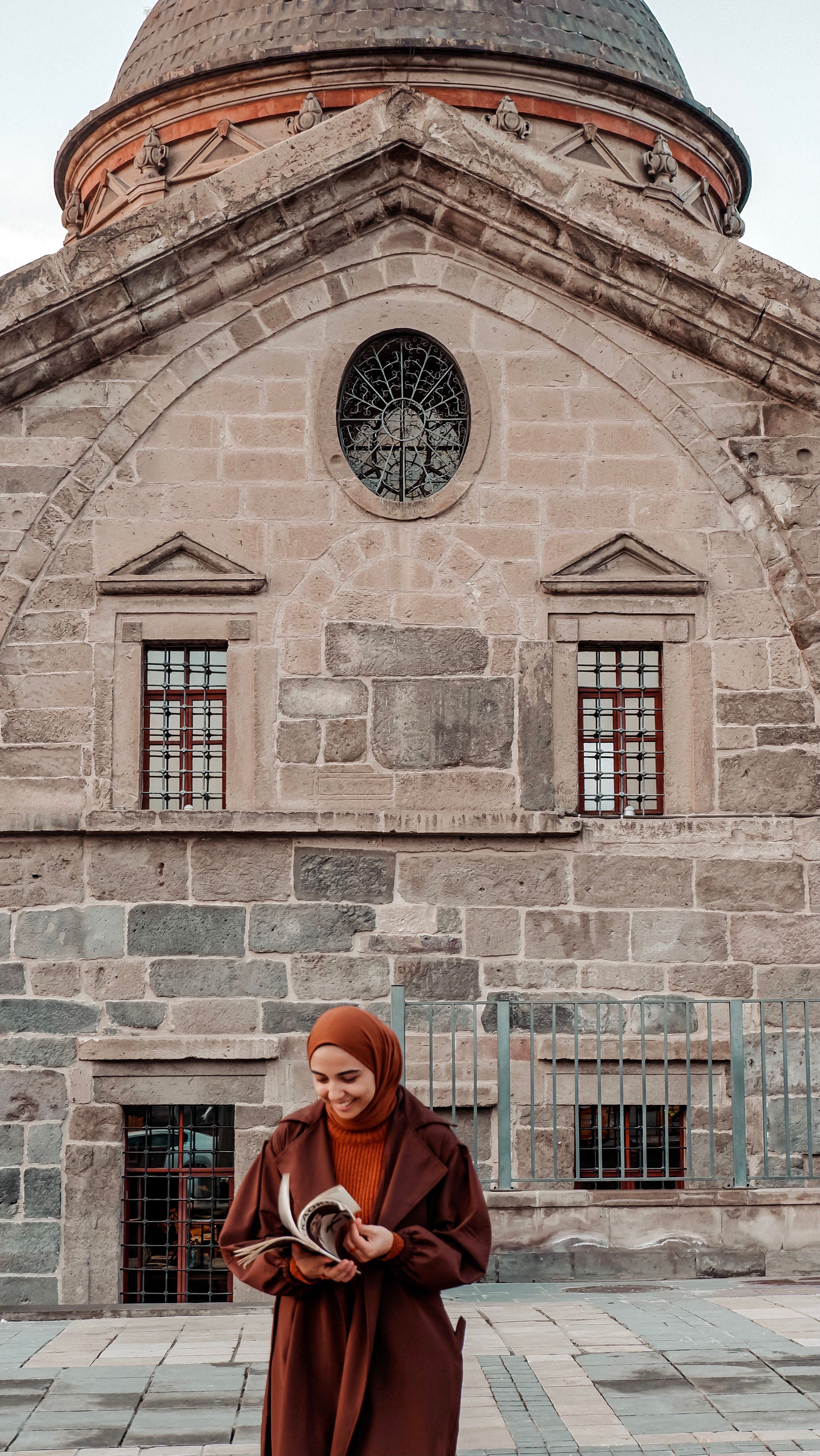 A woman in a hijab stands with a book outside a historic mosque in Kayseri, Türkiye.