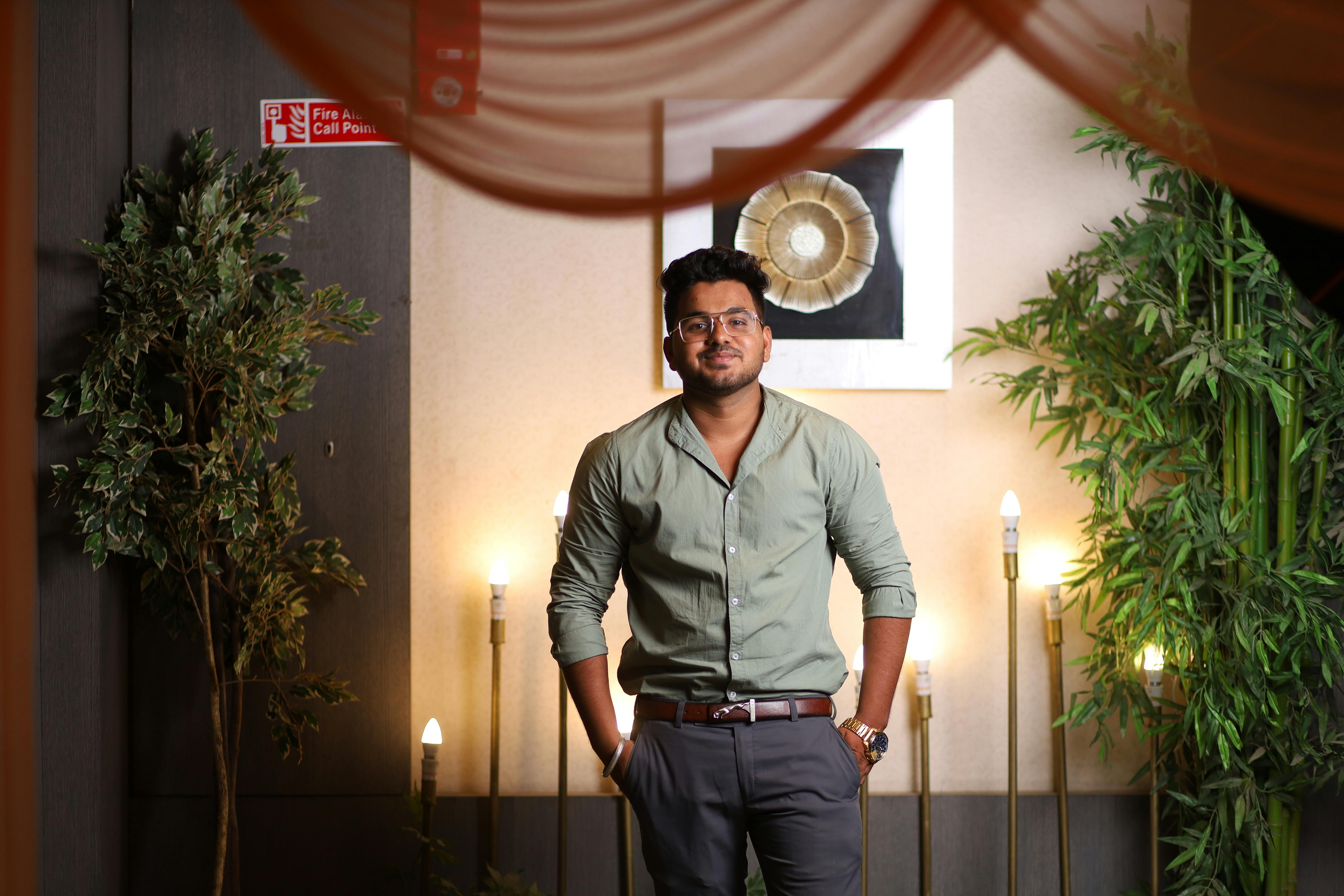 Man wearing green long sleeves, standing confidently indoors, surrounded by decor and lamps.