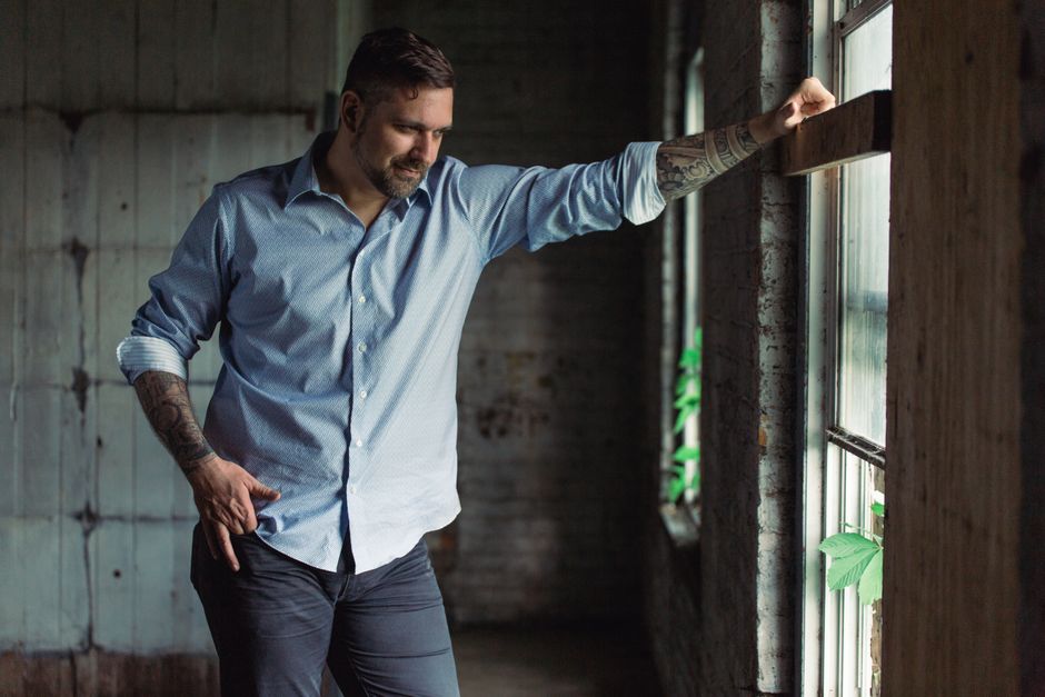 A thoughtful man with tattoos gazes out the window in a stylish shirt, capturing a pensive indoor moment.