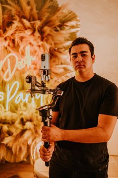 Man standing indoors holding a smartphone gimbal. Neon lights and decor in background.