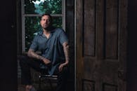 一个纹身男子坐在室内，神情阴郁，窗外是郁郁葱葱的绿植