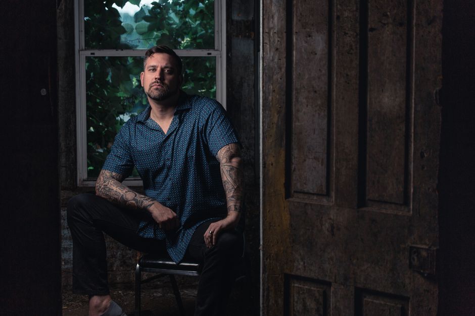 A moody portrait of a tattooed man sitting indoors by a window with lush greenery outside.