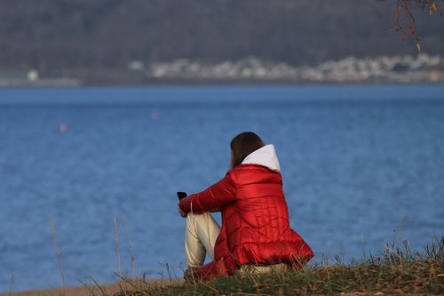 Free Woman in a red jacket sits by Lake Vättern in Jönköping, Sweden, enjoying the peaceful waterside view. Stock Photo