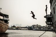 Side view of full length black boy jumped into water in port while friend waiting queue to jump down on abandoned ship against old town in overcast weather